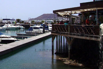 Gemütliches Restaurant an einem Yachthafen auf der kanarischen Insel beim Lanzarote Urlaub. Zu sehen Touristen beim Speisen unter dem Schatten spendenden Vordach auf der Terrasse und im Hintergrund schöne Yachten die vor Anker liegen.